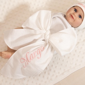 Puede incluir: Una muñeca para bebé envuelta en una manta blanca con textura en forma de lazo, bordada con el nombre "Margo" en rosa. La muñeca lleva un gorro blanco con texto rosa y un body blanco. Descansa sobre una manta blanca con textura.