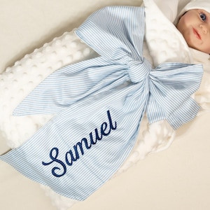 May include: A white baby blanket with a large blue and white striped bow. The name "Samuel" is embroidered in navy blue script on one of the bow's tails. The blanket has a textured, dotted surface.