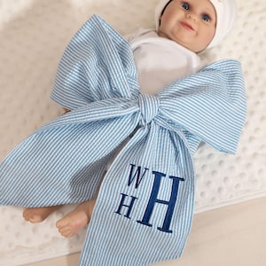 Peut inclure: Une poupée bébé portant un bonnet blanc et un body blanc, enveloppée dans un grand nœud en tissu seersucker bleu clair et blanc. Le nœud présente un monogramme brodé bleu foncé avec les lettres WHH sur l'un des pans longs et pointus.