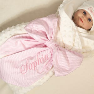 May include: A baby swaddling blanket with a large pink and white striped bow. The blanket is embroidered with the name "Sophia" in pink script. A white hat with the name "Summer" is on the baby's head.