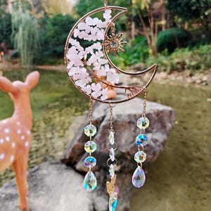 May include: A copper-colored crescent moon suncatcher with a sun charm and dangling iridescent crystals. The moon is filled with pink stones and wire detailing. The suncatcher hangs from a chain.
