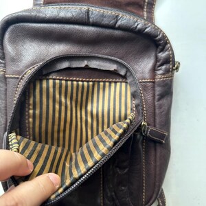 Op de afbeelding: Een donkerbruine leren crossbodytas met een hoofdvak met ritssluiting en een gestreepte binnenkant. De tas heeft een klepsluiting en verstelbare bandjes. De stiksels zijn zichtbaar in een lichtere bruine kleur.