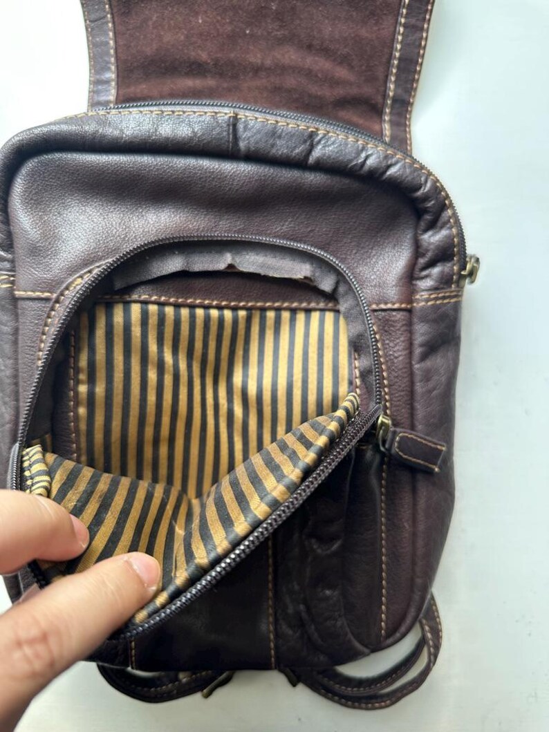 Op de afbeelding: Een donkerbruine leren crossbodytas met een hoofdvak met ritssluiting en een gestreepte binnenkant. De tas heeft een klepsluiting en verstelbare bandjes. De stiksels zijn zichtbaar in een lichtere bruine kleur.