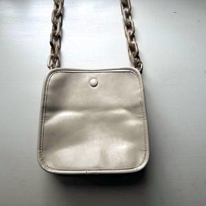 May include: A small, light gray crossbody bag with a square shape and a single button closure. The bag features a chain strap with large, marbled links. The bag is made of a soft, possibly leather-like material.