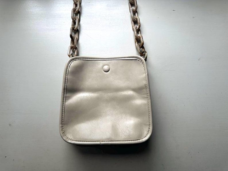May include: A small, light gray crossbody bag with a square shape and a single button closure. The bag features a chain strap with large, marbled links. The bag is made of a soft, possibly leather-like material.