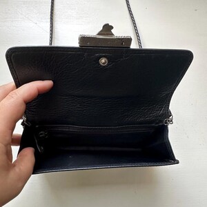 K&ouml;nnte beinhalten: Eine schwarze Ledertasche mit einem silbernen Verschluss und einem Kettenriemen. Die Tasche ist ge&ouml;ffnet und das Innere ist mit schwarzem Stoff ausgekleidet.