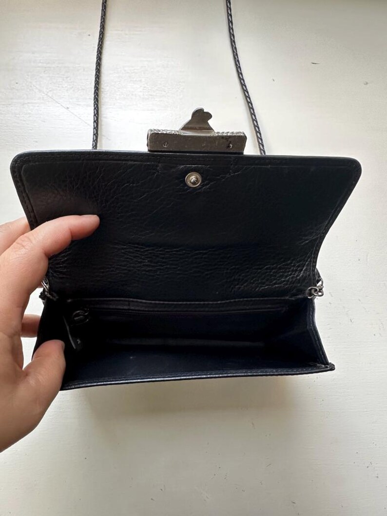 K&ouml;nnte beinhalten: Eine schwarze Ledertasche mit einem silbernen Verschluss und einem Kettenriemen. Die Tasche ist ge&ouml;ffnet und das Innere ist mit schwarzem Stoff ausgekleidet.