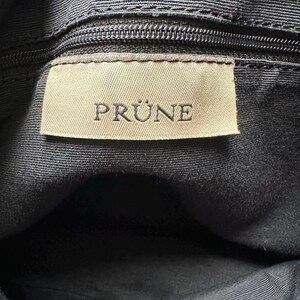 May include: Close-up of a black fabric bag interior with a zipper and a rectangular label. The label is beige with the word "PRUNE" printed in capital letters.