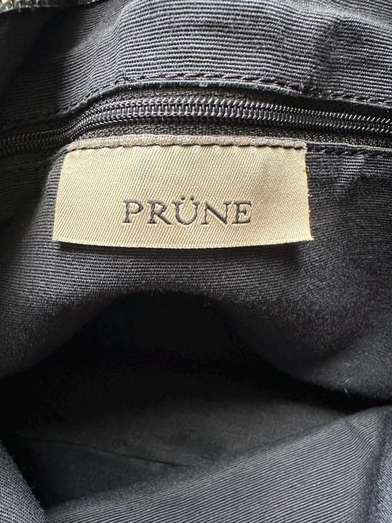 May include: Close-up of a black fabric bag interior with a zipper and a rectangular label. The label is beige with the word "PRUNE" printed in capital letters.