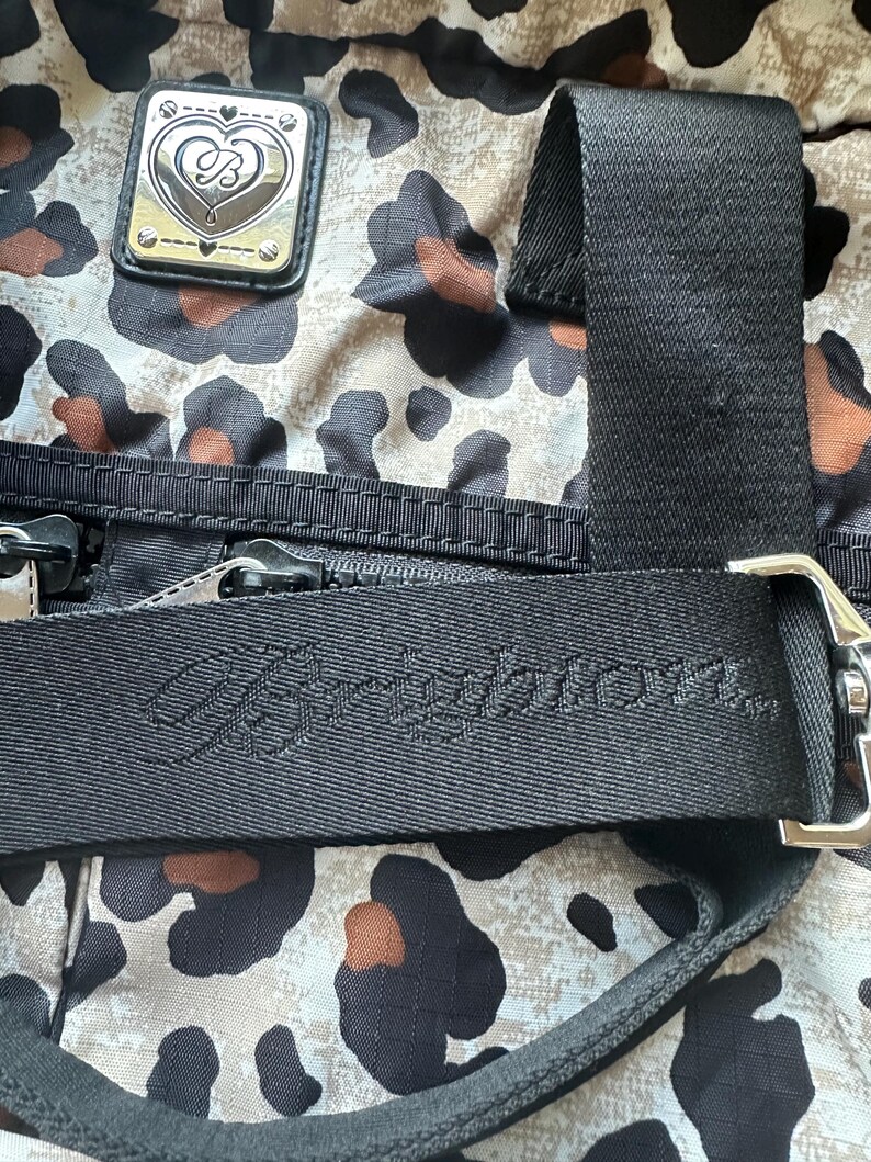 May include: A black and white leopard print bag with a black strap and a silver heart-shaped logo with the text "Juicy Couture" on the strap.