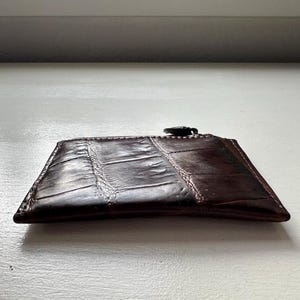 Puede incluir: Un tarjetero de cuero marr&oacute;n oscuro con un patr&oacute;n texturizado similar al de la piel de cocodrilo. El tarjetero tiene forma rectangular y cierre de cremallera en la parte superior. El cuero es de color marr&oacute;n oscuro.