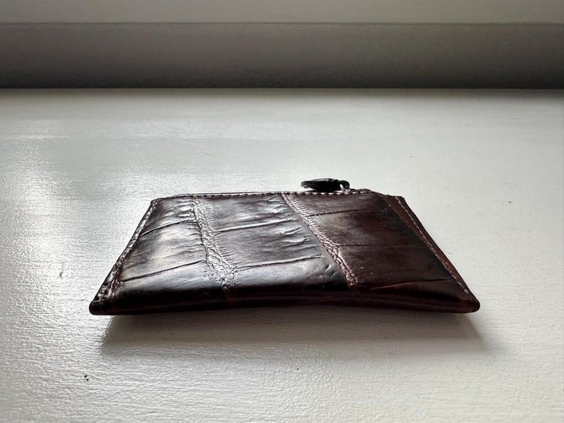 Puede incluir: Un tarjetero de cuero marr&oacute;n oscuro con un patr&oacute;n texturizado similar al de la piel de cocodrilo. El tarjetero tiene forma rectangular y cierre de cremallera en la parte superior. El cuero es de color marr&oacute;n oscuro.