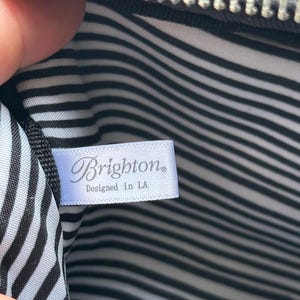 K&ouml;nnte beinhalten: Innenansicht einer schwarzen Tasche mit silbernem Rei&szlig;verschluss. Das Futter weist schwarz-wei&szlig;e horizontale Streifen auf. Ein wei&szlig;es Etikett tr&auml;gt die Aufschrift "Brighton" und "Designed in LA".