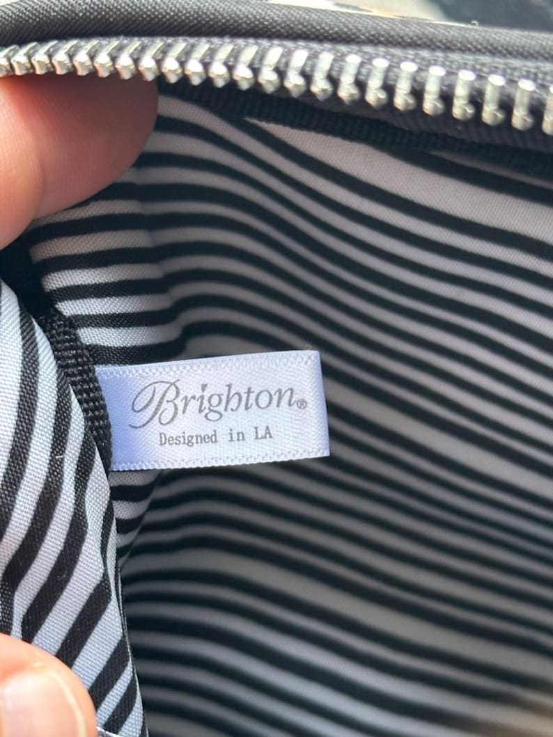 K&ouml;nnte beinhalten: Innenansicht einer schwarzen Tasche mit silbernem Rei&szlig;verschluss. Das Futter weist schwarz-wei&szlig;e horizontale Streifen auf. Ein wei&szlig;es Etikett tr&auml;gt die Aufschrift "Brighton" und "Designed in LA".