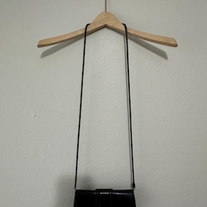 K&ouml;nnte beinhalten: Eine schwarze Leder-Umh&auml;ngetasche mit einem silbernen herzf&ouml;rmigen Verschluss. Die Tasche h&auml;ngt an einem Holzb&uuml;gel.