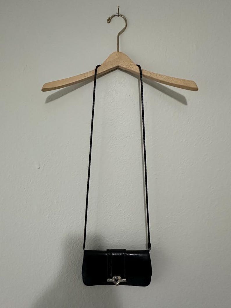 K&ouml;nnte beinhalten: Eine schwarze Leder-Umh&auml;ngetasche mit einem silbernen herzf&ouml;rmigen Verschluss. Die Tasche h&auml;ngt an einem Holzb&uuml;gel.