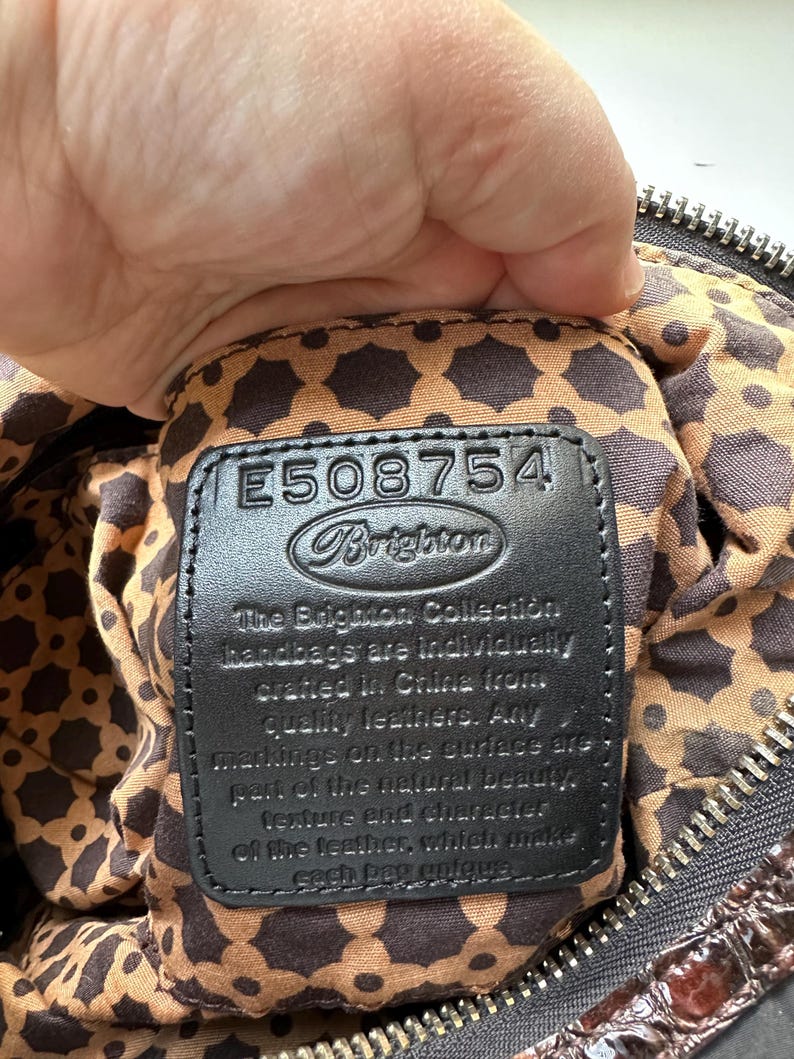 May include: Close-up of a black leather handbag tag with the text "The Brighton Collection handbags are individually crafted in China from quality leathers. Any markings on the surface are part of the natural beauty, texture and character of the leather, which makes each bag unique." and the number "E508754" above the text.