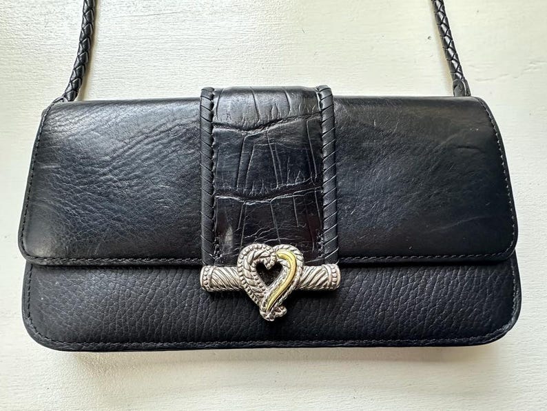 K&ouml;nnte beinhalten: Eine schwarze Lederhandtasche mit einem silbernen herzf&ouml;rmigen Verschluss und einem geflochtenen Riemen. Die Tasche hat ein strukturiertes, krokodilgepr&auml;gtes Panel in der Mitte.