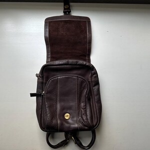 Op de afbeelding: Een donkerbruine leren rugzak met een flap en een klein voorvak. De tas heeft een afgeronde vorm met zichtbare stiksels en een goudkleurige knoopsluiting. De bandjes zijn zwart en de binnenkant is lichtbruin.