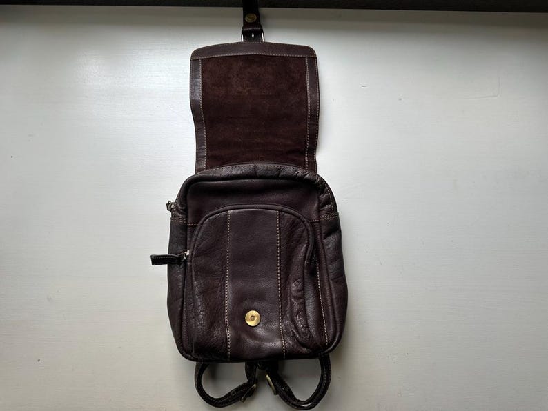 Op de afbeelding: Een donkerbruine leren rugzak met een flap en een klein voorvak. De tas heeft een afgeronde vorm met zichtbare stiksels en een goudkleurige knoopsluiting. De bandjes zijn zwart en de binnenkant is lichtbruin.