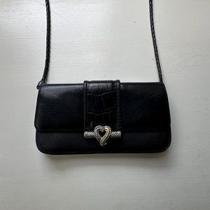 K&ouml;nnte beinhalten: Eine schwarze Leder-Umh&auml;ngetasche mit einem silbernen herzf&ouml;rmigen Verschluss. Die Tasche hat ein strukturiertes Krokodilmuster auf der Vorderseite.