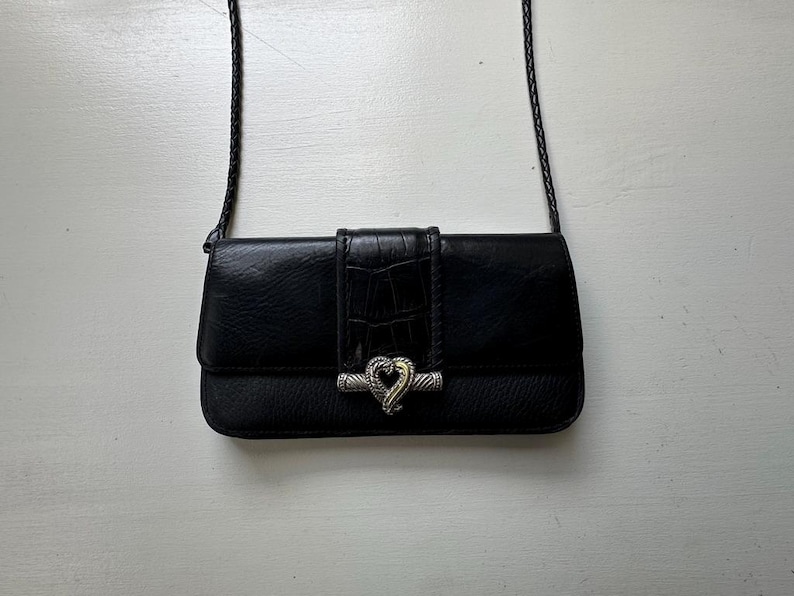 K&ouml;nnte beinhalten: Eine schwarze Leder-Umh&auml;ngetasche mit einem silbernen herzf&ouml;rmigen Verschluss. Die Tasche hat ein strukturiertes Krokodilmuster auf der Vorderseite.