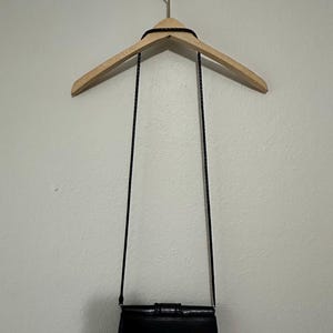 K&ouml;nnte beinhalten: Eine schwarze Leder-Umh&auml;ngetasche mit Klappenverschluss und einem dekorativen Schleifendetail. Die Tasche h&auml;ngt an einem Holzb&uuml;gel.