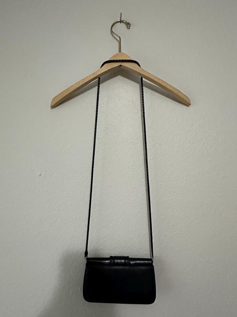 K&ouml;nnte beinhalten: Eine schwarze Leder-Umh&auml;ngetasche mit Klappenverschluss und einem dekorativen Schleifendetail. Die Tasche h&auml;ngt an einem Holzb&uuml;gel.