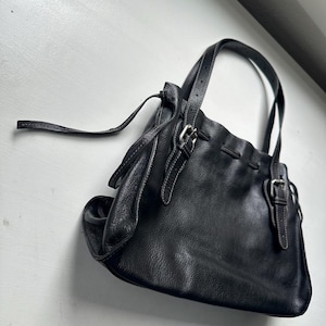 May include: Black leather handbag with two handles and a shoulder strap. The bag features silver-colored buckles and white stitching details. The bag has a soft, slouchy design.
