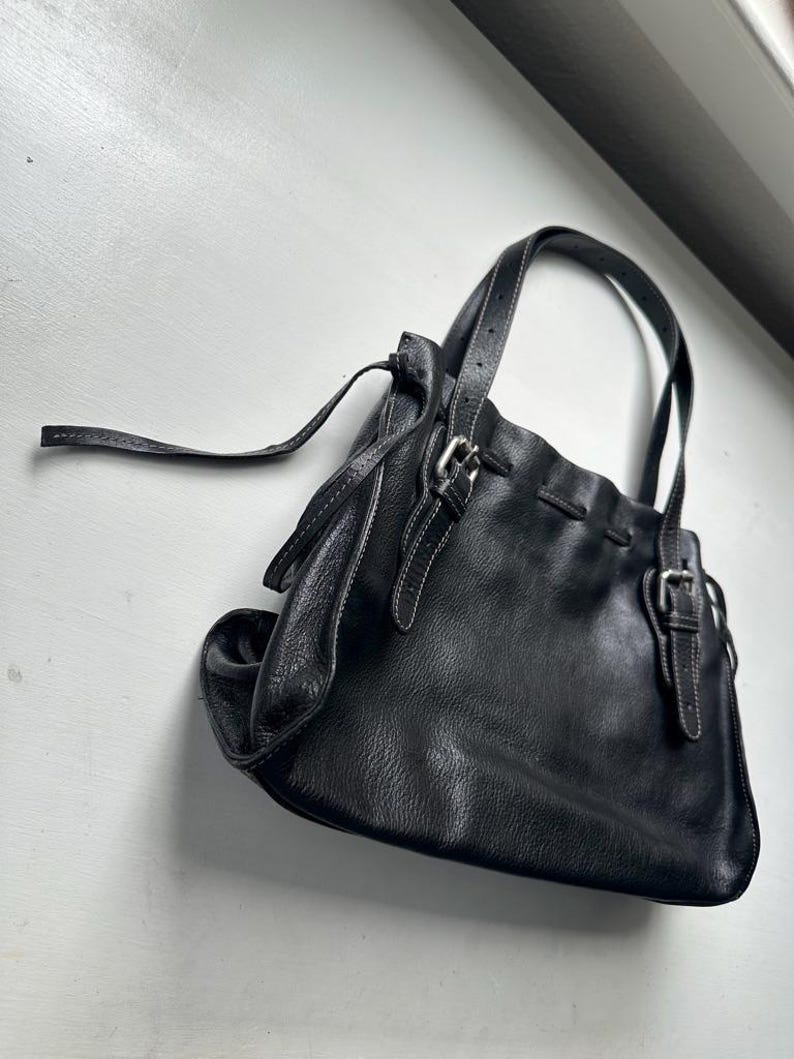 May include: Black leather handbag with two handles and a shoulder strap. The bag features silver-colored buckles and white stitching details. The bag has a soft, slouchy design.