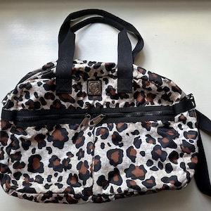 May include: A leopard print travel bag with black straps and a zippered pocket. The bag has a black and silver logo on the front.