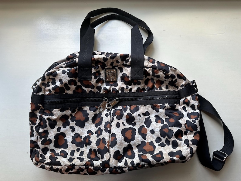 May include: A leopard print travel bag with black straps and a zippered pocket. The bag has a black and silver logo on the front.