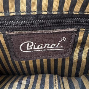 Op de afbeelding: Binnenaanzicht van een tas met een bruin leren label met de merknaam "Bianci" in witte letters. De binnenvoering heeft een gestreept patroon in zwart en goud. Een zwarte ritssluiting is zichtbaar.