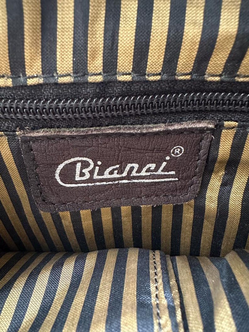 Op de afbeelding: Binnenaanzicht van een tas met een bruin leren label met de merknaam "Bianci" in witte letters. De binnenvoering heeft een gestreept patroon in zwart en goud. Een zwarte ritssluiting is zichtbaar.