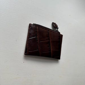 Puede incluir: Un peque&ntilde;o monedero de cuero marr&oacute;n oscuro con un patr&oacute;n texturizado similar al de un cocodrilo. El monedero tiene un cierre de cremallera y una peque&ntilde;a leng&uuml;eta redonda. El monedero es rectangular y parece vac&iacute;o.