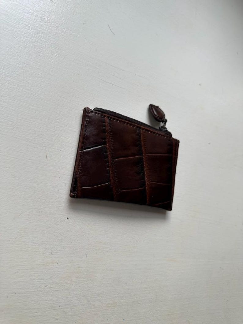 Puede incluir: Un peque&ntilde;o monedero de cuero marr&oacute;n oscuro con un patr&oacute;n texturizado similar al de un cocodrilo. El monedero tiene un cierre de cremallera y una peque&ntilde;a leng&uuml;eta redonda. El monedero es rectangular y parece vac&iacute;o.