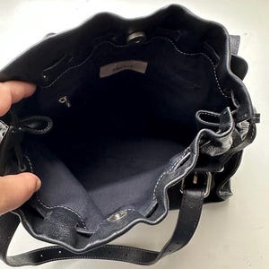 May include: Black leather bucket bag with white stitching and a drawstring closure. The bag has a shoulder strap and a silver-tone clasp. The interior is dark blue, and a small tag reads "PRONE".