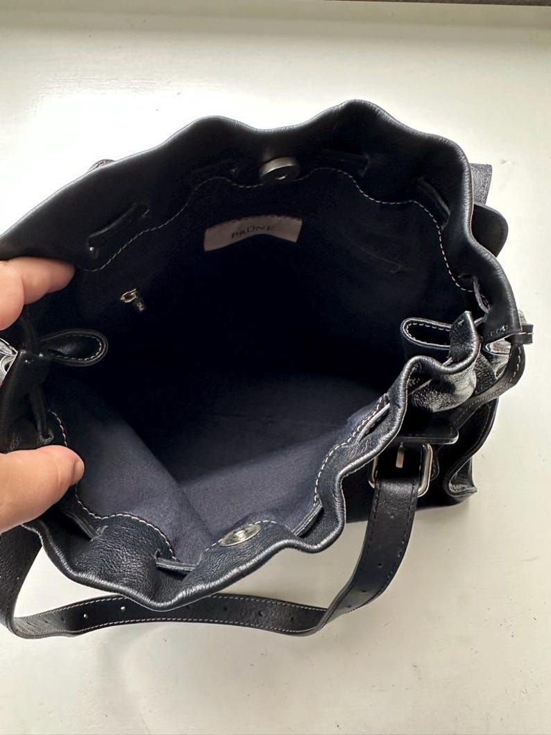 May include: Black leather bucket bag with white stitching and a drawstring closure. The bag has a shoulder strap and a silver-tone clasp. The interior is dark blue, and a small tag reads "PRONE".
