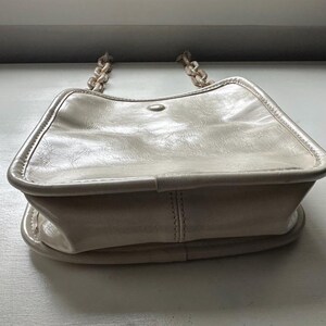May include: Cream-colored handbag with a chain strap. The bag has a rectangular shape with rounded edges and a flap closure. The chain strap is made of a matching cream-colored material. The bag is made of a smooth, possibly leather-like material.