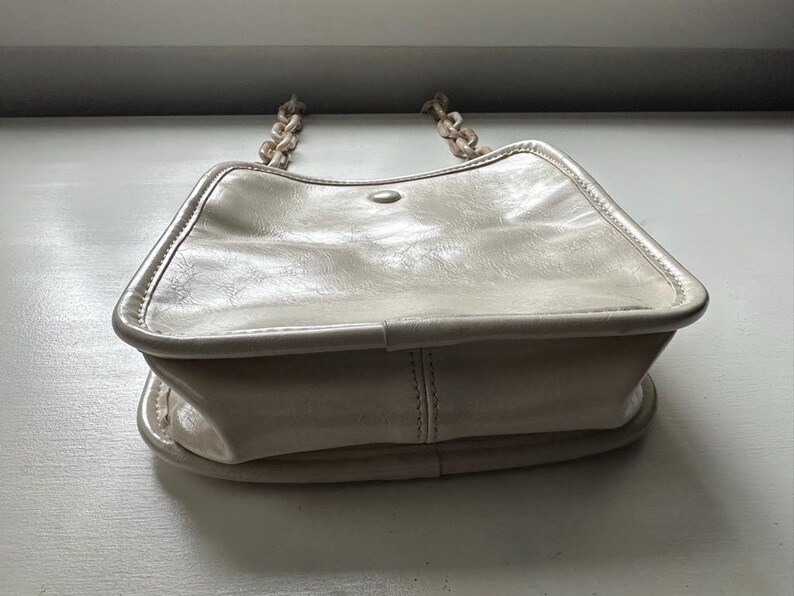 May include: Cream-colored handbag with a chain strap. The bag has a rectangular shape with rounded edges and a flap closure. The chain strap is made of a matching cream-colored material. The bag is made of a smooth, possibly leather-like material.