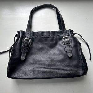 May include: Black leather tote bag with two handles and two buckle closures. The bag has a gathered top and a textured surface. The handles are attached with silver-colored hardware. The bag is sitting on a white surface.