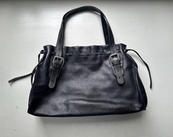 May include: Black leather tote bag with two handles and two buckle closures. The bag has a gathered top and a textured surface. The handles are attached with silver-colored hardware. The bag is sitting on a white surface.