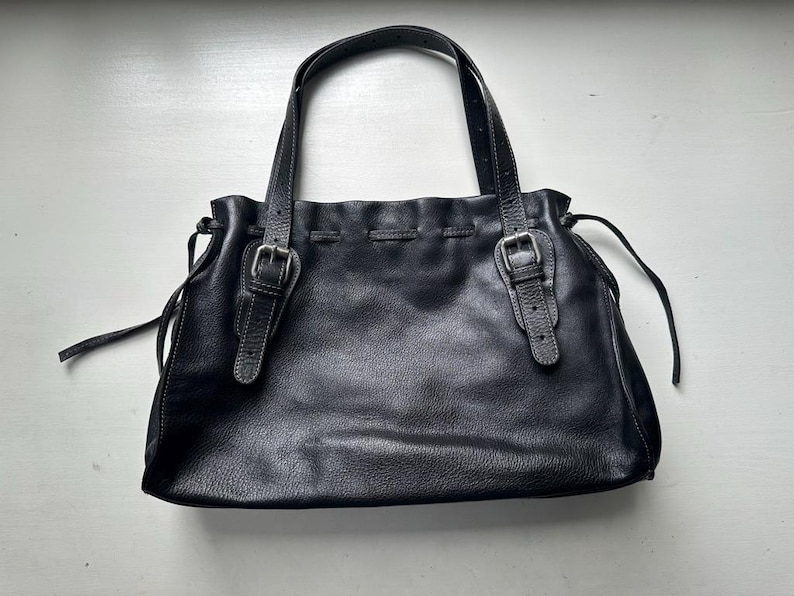 May include: Black leather tote bag with two handles and two buckle closures. The bag has a gathered top and a textured surface. The handles are attached with silver-colored hardware. The bag is sitting on a white surface.