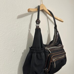 May include: A black nylon handbag with brown faux leather accents and a braided strap. The bag has multiple zippered compartments and a drawstring closure.