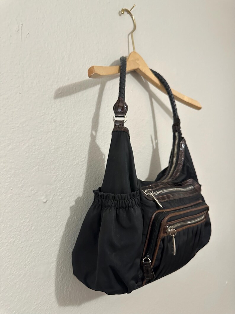 May include: A black nylon handbag with brown faux leather accents and a braided strap. The bag has multiple zippered compartments and a drawstring closure.
