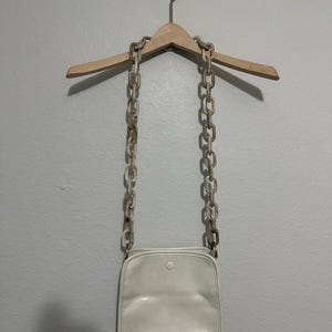 May include: A cream-colored crossbody bag with a square shape and a single button closure. The bag hangs from a light-colored chain strap. The chain is made of large, marbled links. The bag is suspended from a wooden hanger against a gray wall.