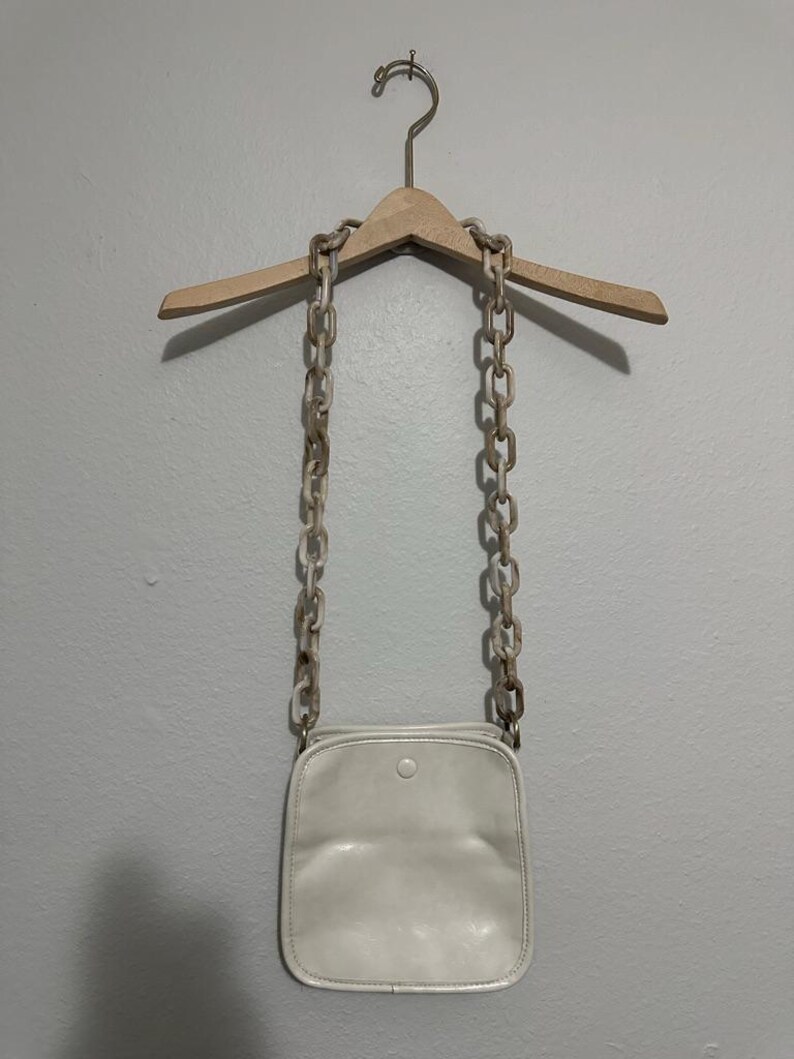 May include: A cream-colored crossbody bag with a square shape and a single button closure. The bag hangs from a light-colored chain strap. The chain is made of large, marbled links. The bag is suspended from a wooden hanger against a gray wall.