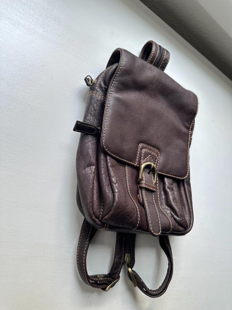 Op de afbeelding: Een kleine, bruine leren rugzak met een klepsluiting en een gesp. De rugzak heeft verstelbare bandjes en een handvat aan de bovenkant. Witte stiksels accentueren de randen. De hardware is goudkleurig.
