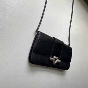 K&ouml;nnte beinhalten: Eine schwarze Leder-Umh&auml;ngetasche mit einem silbernen Schlangenverschluss. Die Tasche hat einen geflochtenen Riemen und einen Klappenverschluss.