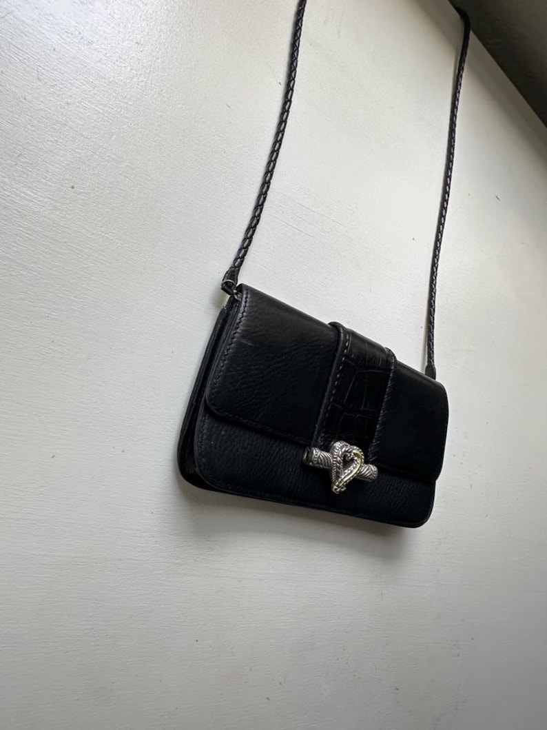 K&ouml;nnte beinhalten: Eine schwarze Leder-Umh&auml;ngetasche mit einem silbernen Schlangenverschluss. Die Tasche hat einen geflochtenen Riemen und einen Klappenverschluss.